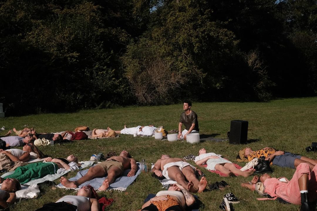 Group breathwork session on grass with participants lying on mats and a facilitator seated with singing bowls